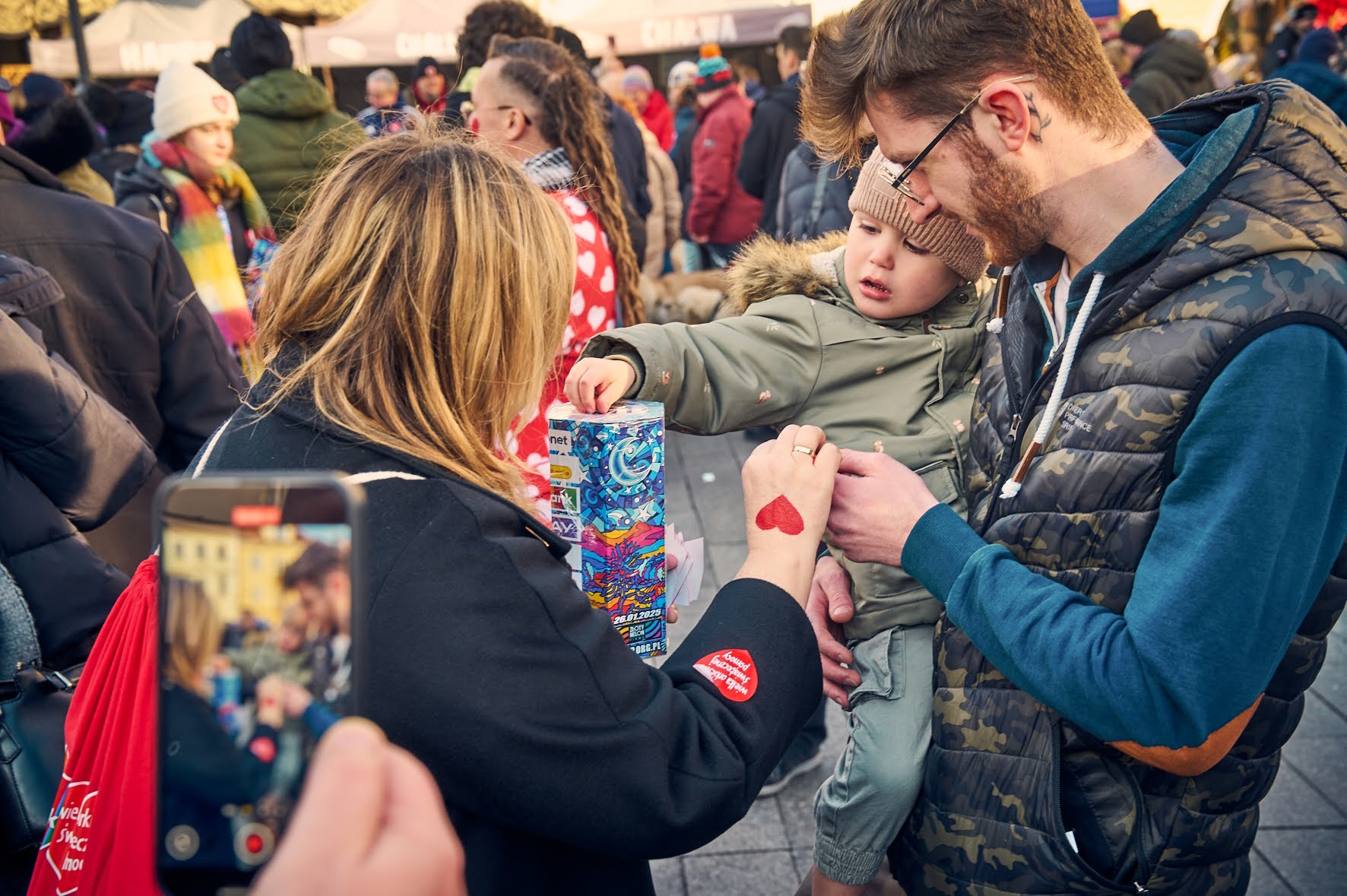 Mężczyzna z chłopcem na rękach, chłopiec wrzuca pieniądze do puszki, którą trzyma prezydent Kuczyńska-Budka